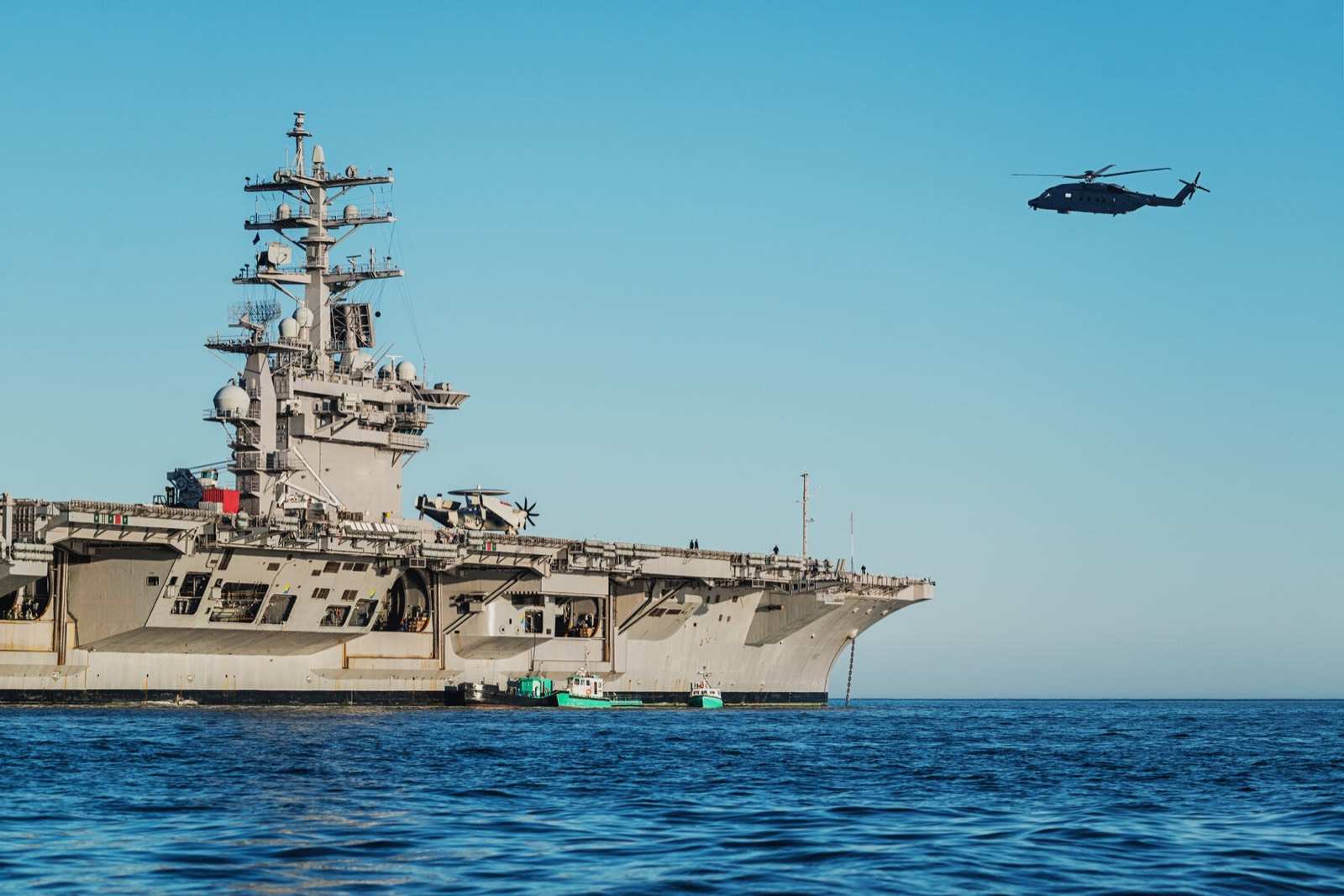 An aircraft carrier under way at sea, a helicopter overhead.